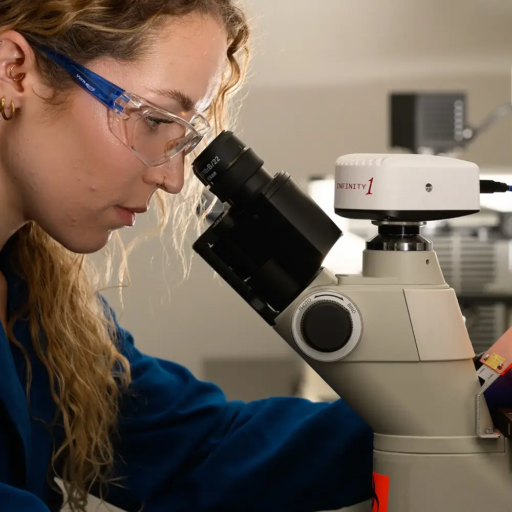 A young woman with long curly hair and safety goggles looks through a microscope in a lab, wearing a blue lab coat.