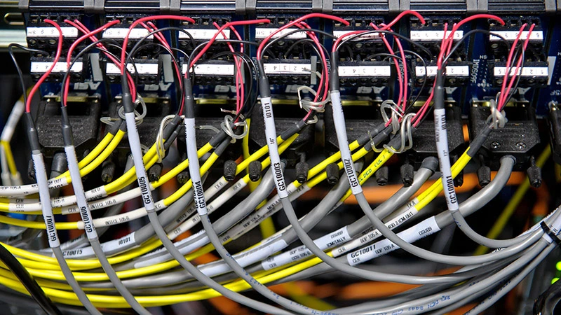 Close-up of densely packed yellow, gray, red, and black cables connecting to dark network hardware. Many cables feature white alphanumeric labels, showing an intricate and organized IT system.
