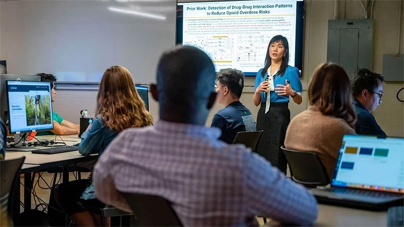 An Asian woman in a blue blouse gestures while presenting research on opioid overdose risks to students in a classroom. Students seated at computers listen or work independently.