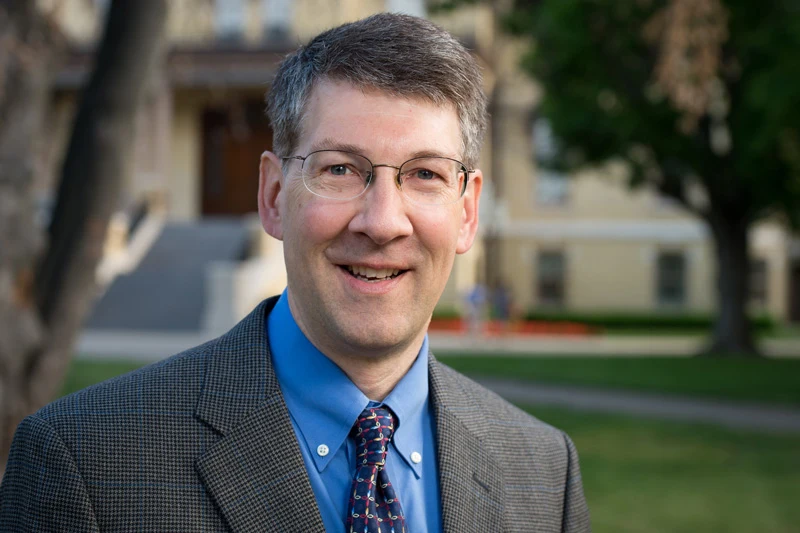 Headshot of a smiling man with glasses, wearing a gray plaid jacket, blue shirt, and patterned tie, outdoors.