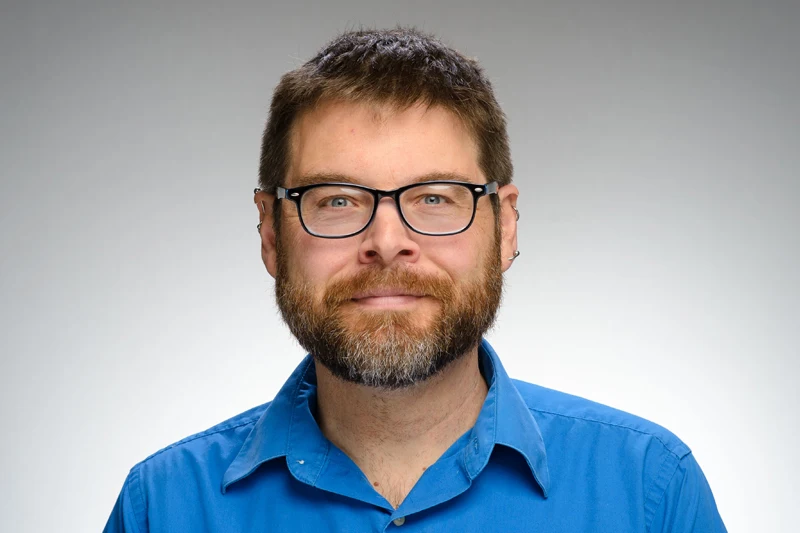 Headshot of a smiling man with a beard and glasses, wearing a blue button-down shirt.