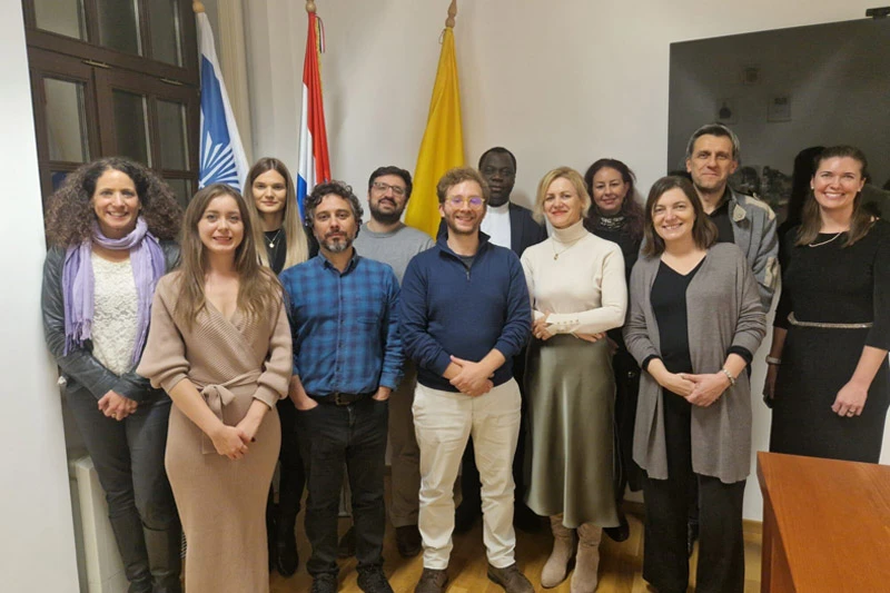 Thirteen smiling men and women, diverse in appearance, stand indoors in business casual attire. Behind them are the European Union flag, the Croatian flag, and a yellow flag.