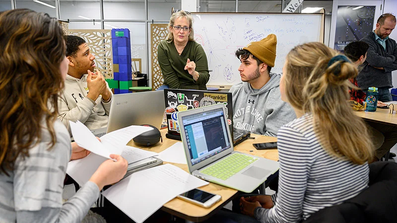 A female instructor in a green shirt gestures while speaking to four students with laptops and papers at a table in a bright classroom. 