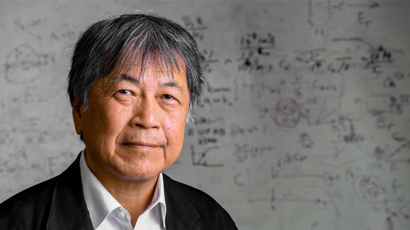 An older Asian man with graying hair smiles gently, wearing a black jacket and white shirt. Behind him is a whiteboard covered in blurry scientific equations and diagrams.