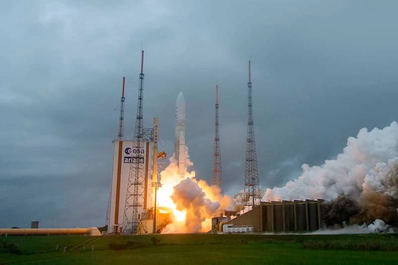An Ariane rocket launches, with bright orange flames and white smoke billowing from the base.  The rocket is white and tall, ascending against a cloudy gray sky.  Three tall metal towers surround the launchpad.
