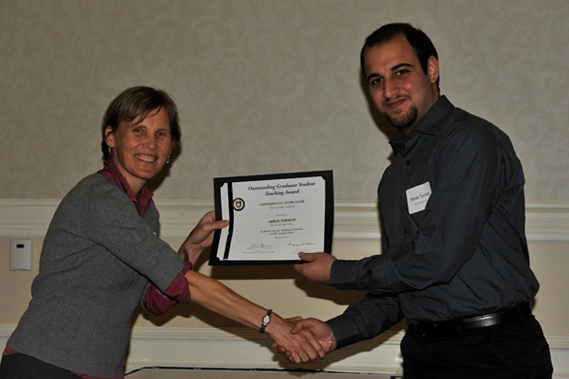 A smiling woman presents a certificate for the Outstanding Graduate Student Teaching Award to a man in a dark shirt as they shake hands.