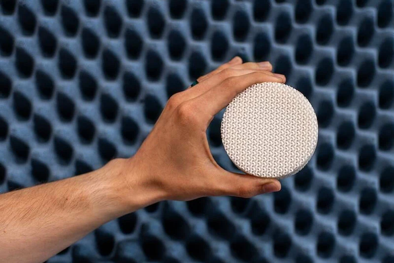 A hand holds a round, white, 3D-printed disc with a woven pattern against a dark blue, acoustically treated, egg-crate-like wall.