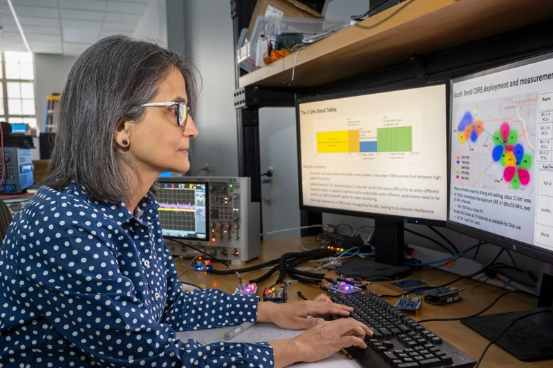 Woman with dark hair and glasses wears a blue polka-dot shirt while typing at a desk with two computer monitors displaying data.