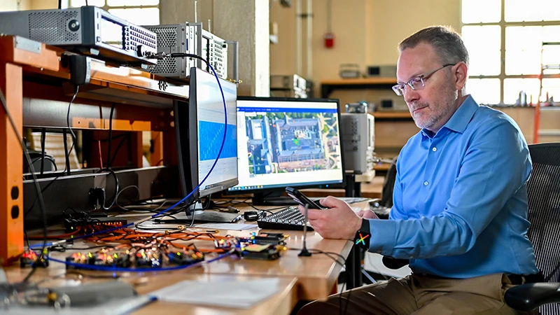 A focused man in a blue shirt and glasses sits at a lab desk, holding a smartphone. The desk is covered in circuit boards, wires, and electronic devices. Two monitors display a blue waveform and an aerial map of a campus building.